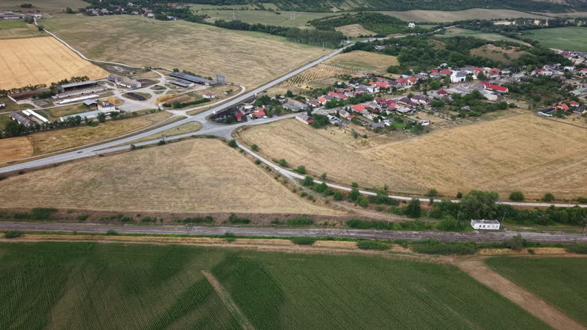 An aerial drone view shows a rural village, vast agricultural fields, intersecting roads, and railway tracks under a summer sky.