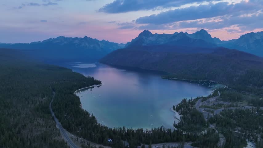 Aerial view of Redfish Lake surrounded by dense pine forests and the Sawtooth Mountains under a twilight sky in Stanley, Idaho, United States.