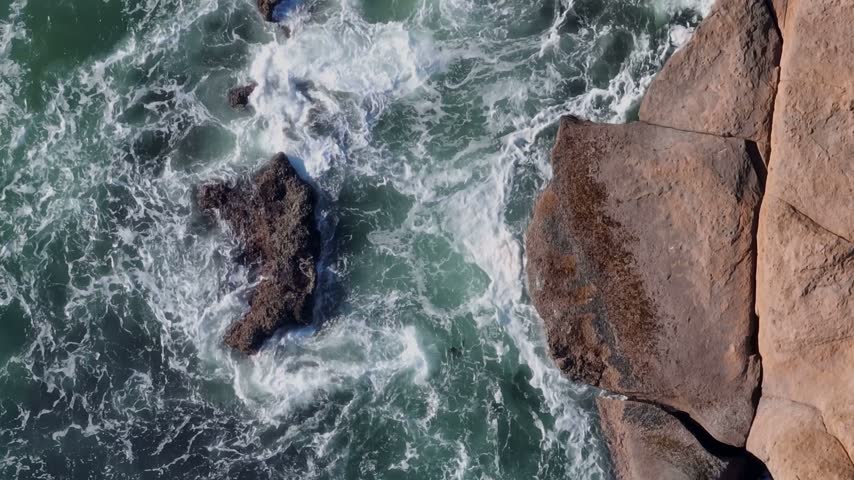 Aerial view of ocean waves crashing against rugged coastal rocks with white sea foam and deep blue water in Oregon City, Oregon, United States.