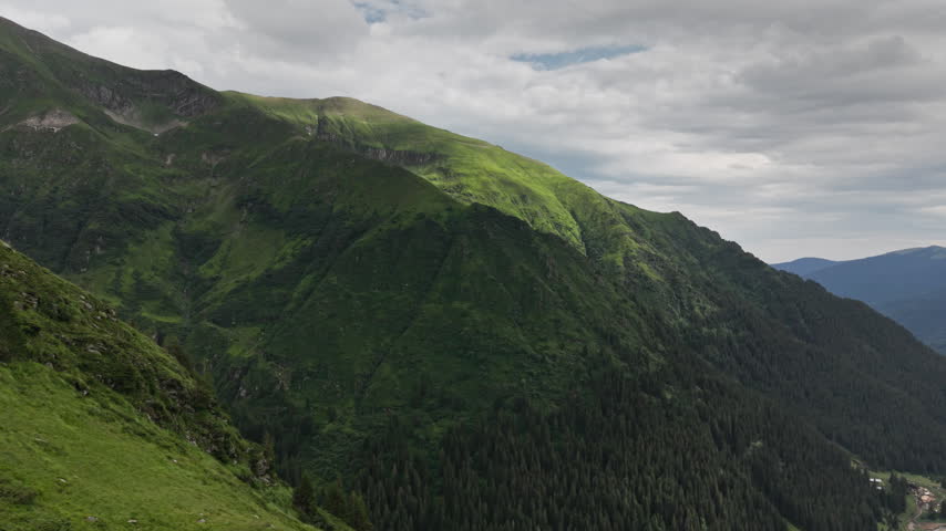 Dramatic green mountain landscape along Romania Transfagarasan route, with steep Carpathian slopes, forested valleys, and clouds overhead.
