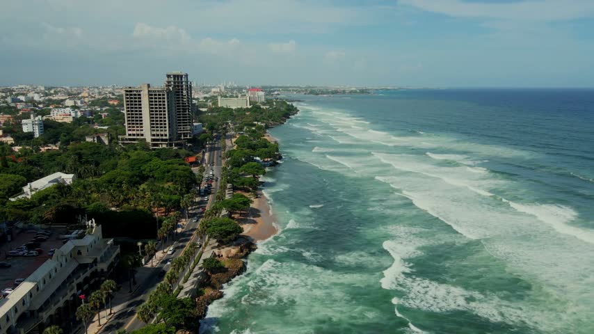 Aerial view of the coastline of Santo Domingo with crashing waves, lush green trees, and urban buildings along the shore in Santo Domingo, Distrito Nacional, Dominican Republic.