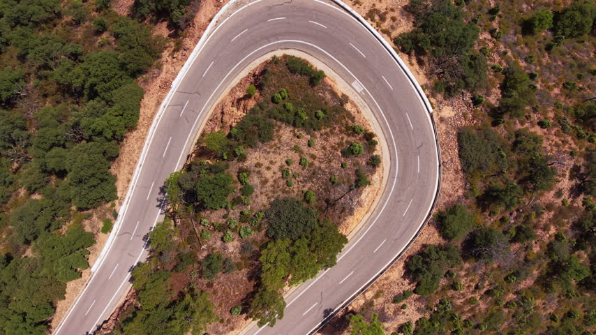 Overhead top-down view of a sharp hairpin turn on a winding mountain road, a white car navigating the curve surrounded by Mediterranean scrubland and green forest