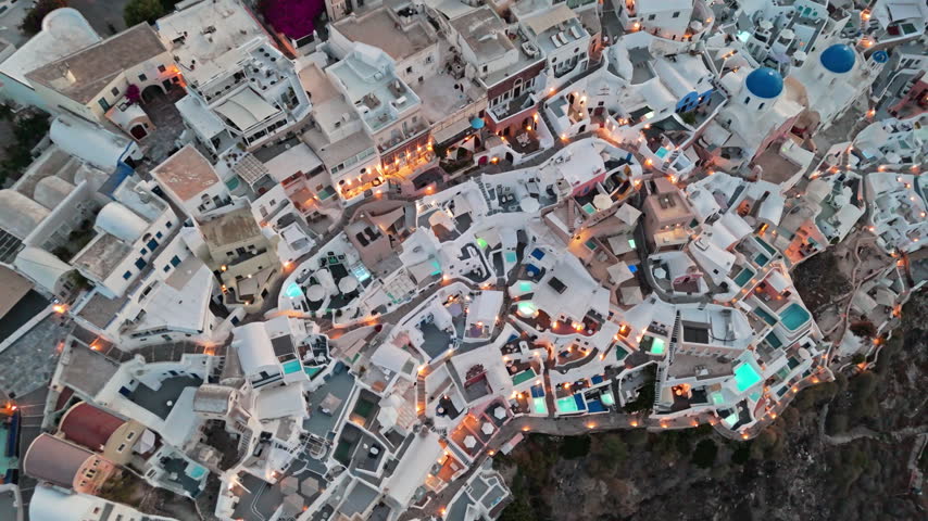Dense cluster of cave-style hotels, rooftop patios, and glowing pools in Oia Santorini as a drone moves slowly overhead at sunrise.