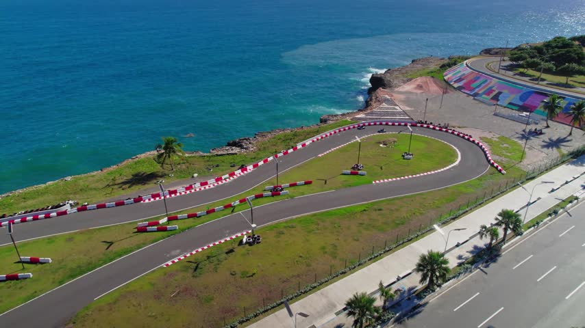 High-angle aerial drone shot pulling back and ascending over a seaside go-kart track, revealing racing karts, the busy coastal highway, and the turquoise Caribbean Sea