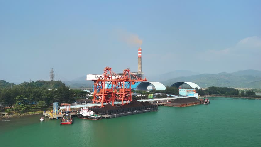 Aerial view of a coal power plant with a tall chimney and large red cranes loading coal onto barges at a coastal terminal