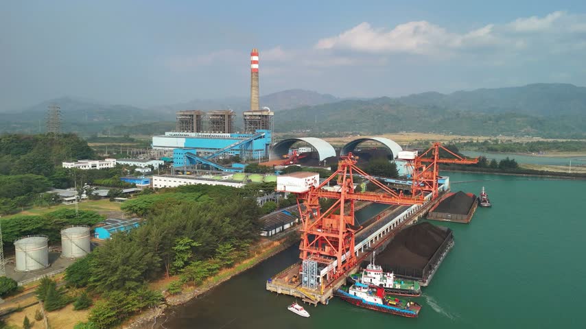 Aerial view of a coal power plant with a tall chimney, large coal storage piles, and red loading cranes at a dock with barges in the water