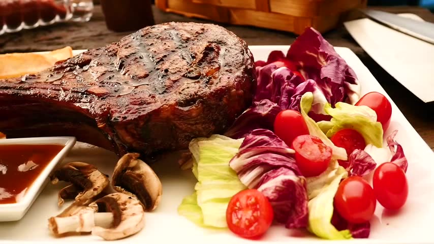 Grilled tomahawk steak served on a white plate with fresh mixed salad, french fries, mushrooms, and dipping sauce. Delicious gourmet meat dish.
