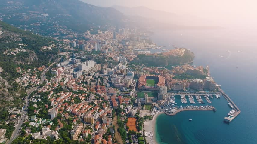Air views of Monaco capture the cityscape with buildings, harbor, and coastline. Boats are seen in the water while the sun sets over the hills in the background.