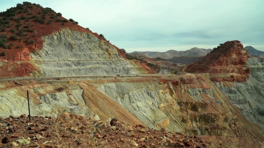 Hillside showcasing distinct layers of colorful rock formations and steep slopes under a cloudy sky. Dirt road winds through the rugged mountain landscape capturing natural beauty.