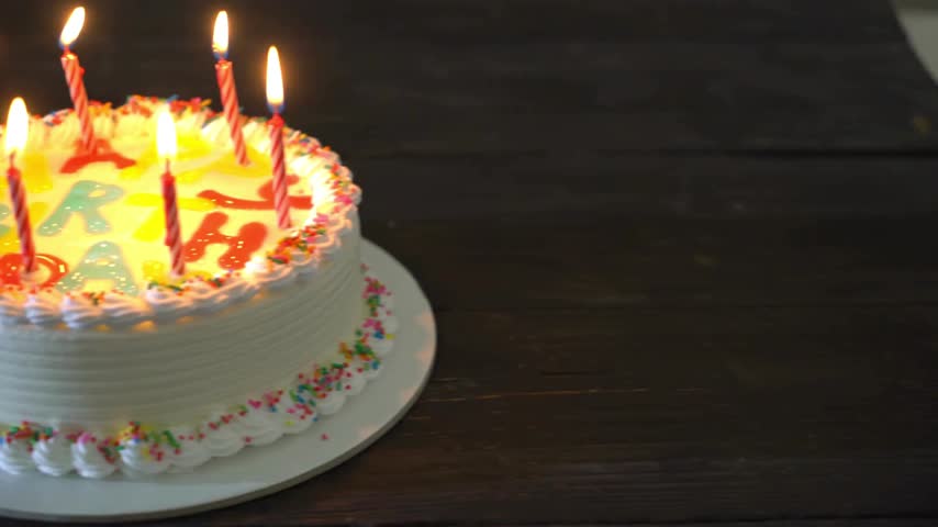 Cake sits on a round platter with colorful candles lighting up the surface. The background features a wooden table. Bright colors create a fun atmosphere for celebration.