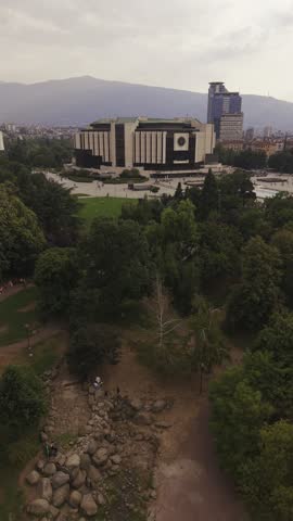 SOFIA, BULGARIA – AUGUST 18, 2026: Wide aerial drone view of central Sofia from the National Palace of Culture area, showing residential buildings, urban greenery, fountains and Vitosha Mountain on the horizon