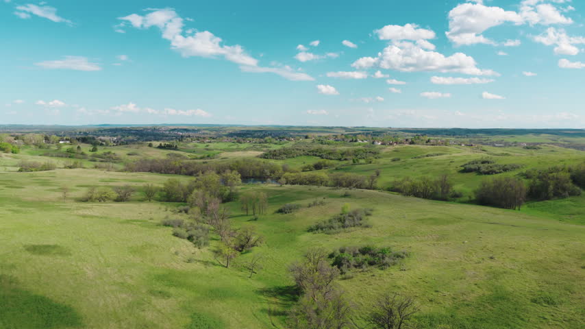 Aerial view over green prairie grassland and rolling hills near Mandan, North Dakota, with a small pond, brushy draws and scattered trees under a bright spring sky.