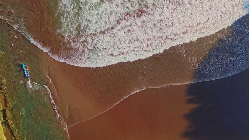 Aerial view of a small boat on the sandy shore of Baron Beach with crashing waves in Yogyakarta, Special Region of Yogyakarta, Indonesia.