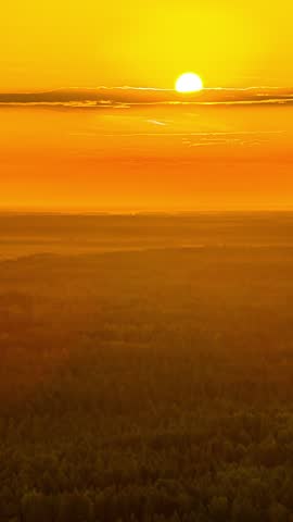 Bright golden sun above hazy horizon over forest at sunrise, aerial vertical hazy glow warming across tree canopy, backdrop