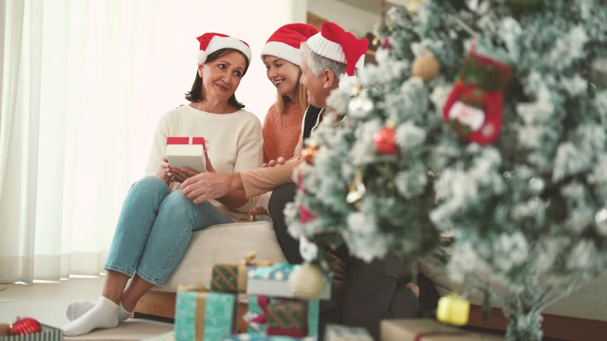Elderly woman receiving Christmas gift from senior man while daughter shares the moment, holiday surprise, family bonding, gift giving, Christmas family celebration.