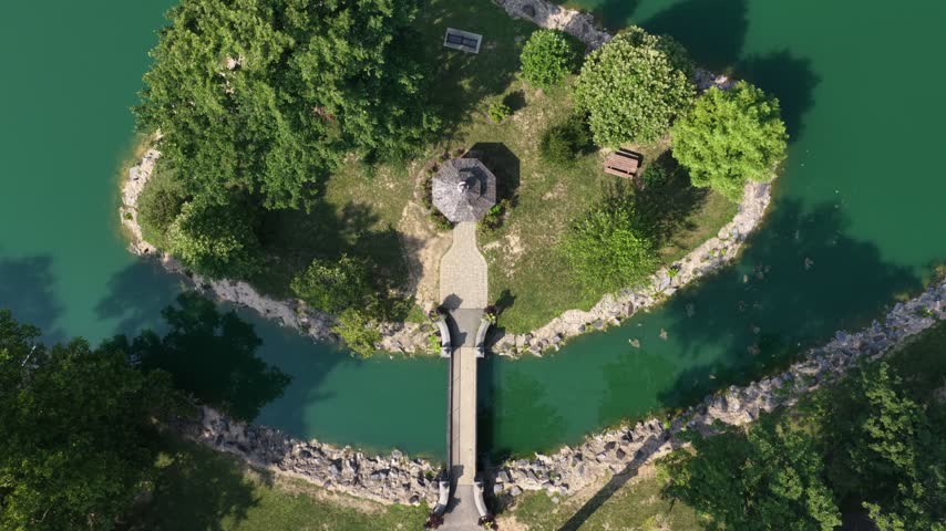 Octagonal gazebo sits center of small wooded island linked to shore by wooden bridge, benches scattered among trees, teal green lake surrounds Longs Park in Pennsylvania. Aerial drone top down shot