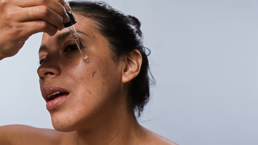 Closeup shot of young Hispanic woman applying cosmetic serum with dropper on face and massaging skin. Clip is ideal for skincare product marketing and beauty e-commerce campaigns