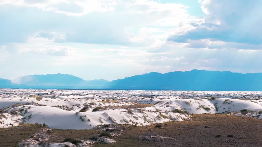 Aerial drone perspective over White Sands National Park in New Mexico, highlighting vast white gypsum sand dunes, desert expanse, and distant mountains under a dramatic sky with clouds