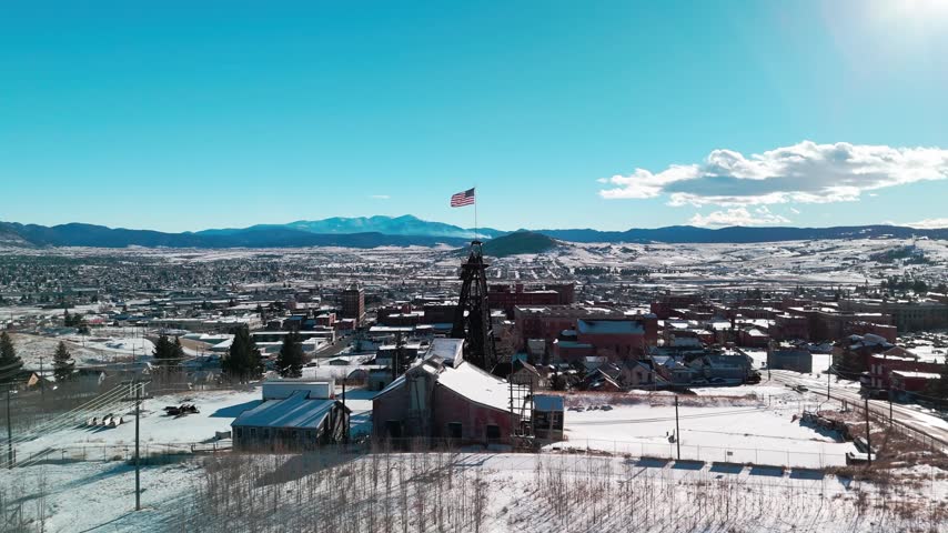Winter aerial drone view over Butte, Montana, capturing snow-covered roofs of historic mining buildings and dense urban architecture extending toward the surrounding regional mountain