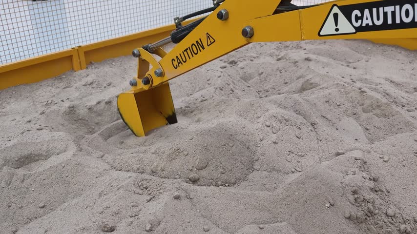 A close-up shot of a yellow miniature toy excavator arm digging into loose sand. The mechanical digger with caution stickers operates inside an enclosed sandbox play area under bright diffuse lighting