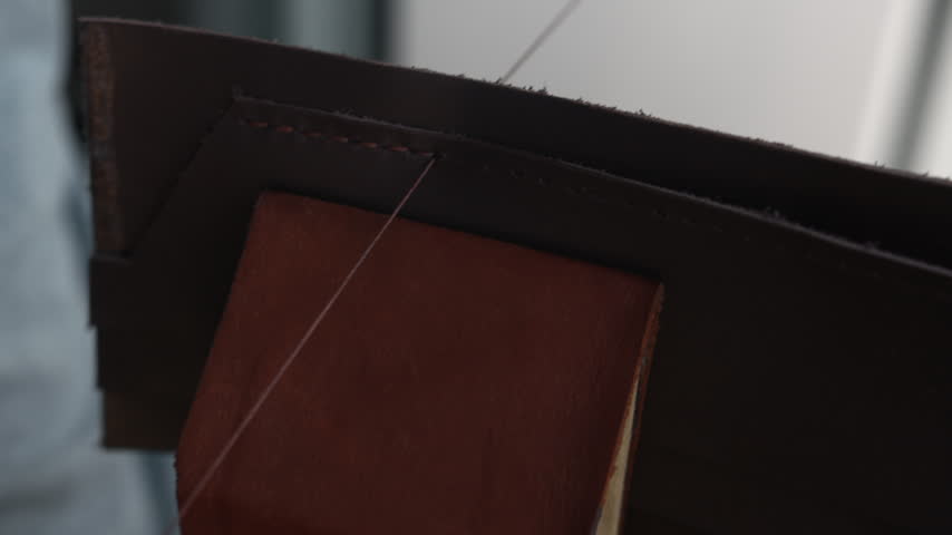 Leather craftsman hands hand stitching a genuine leather wallet with a needle and thread in a workshop, close up
