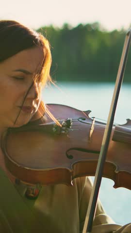 Focused female violinist performing by tranquil lakeside at sunset, graceful musician in flowing dress playing soulful melody in peaceful natural landscape