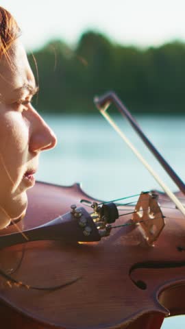 Peaceful closeup portrait of a focused violinist playing by a tranquil lakeside at golden hour, with warm sunlight illuminating her thoughtful expression