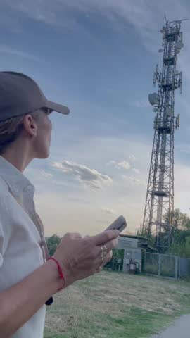 A woman wearing a cap interacts with her smartphone near a large communication tower, highlighting modern connectivity and the widespread reach of mobile technology in an outdoor setting at dusk.