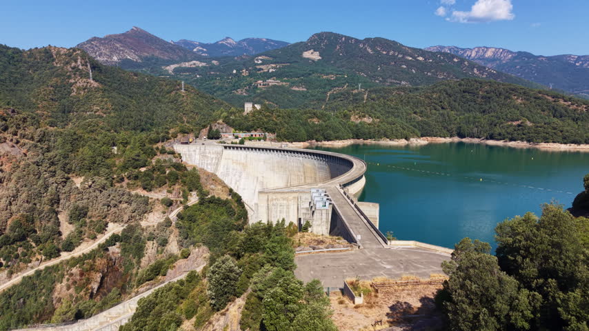 Aerial view of a massive concrete arch dam holding back a blue-green reservoir nestled among forested mountains under a clear summer sky