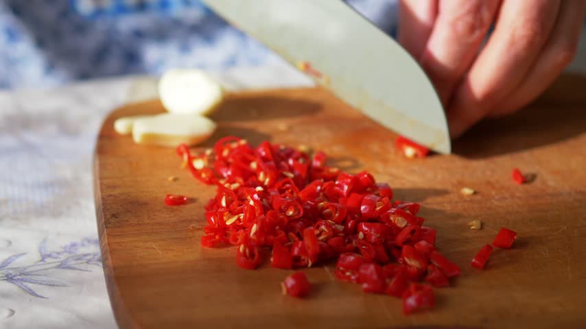 Hot red chili rings and garlic cloves being sliced on rustic wooden board in natural sunlight