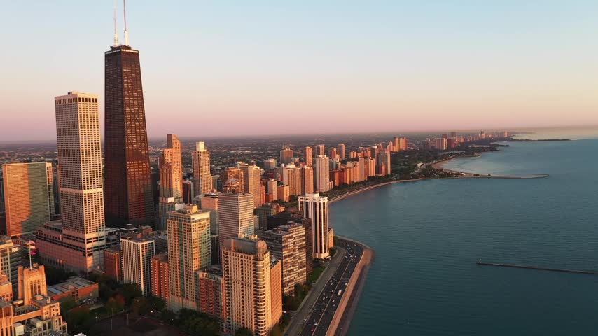 Aerial view of the Chicago lakefront in the morning, with skyscrapers and residential buildings illuminated by warm early sunlight beside the blue waters of Lake Michigan.