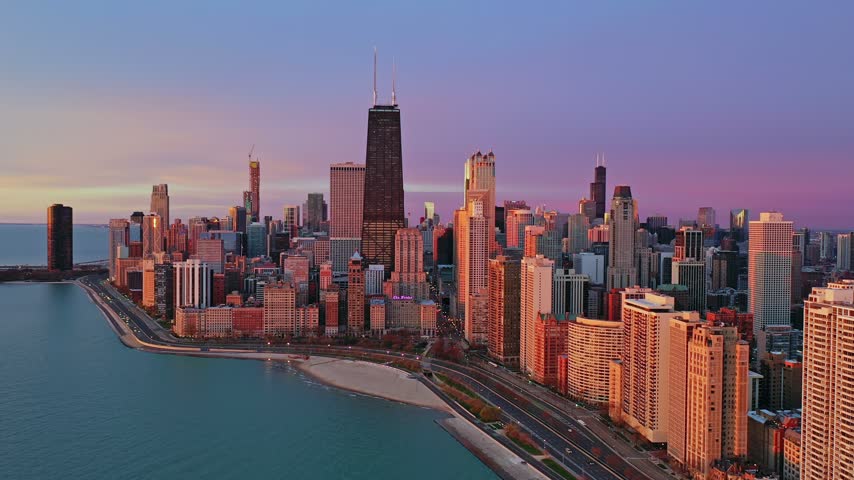 Aerial view of the Chicago skyline in the morning, with downtown skyscrapers glowing in warm early sunlight along the Lake Michigan waterfront and lakefront roadway.