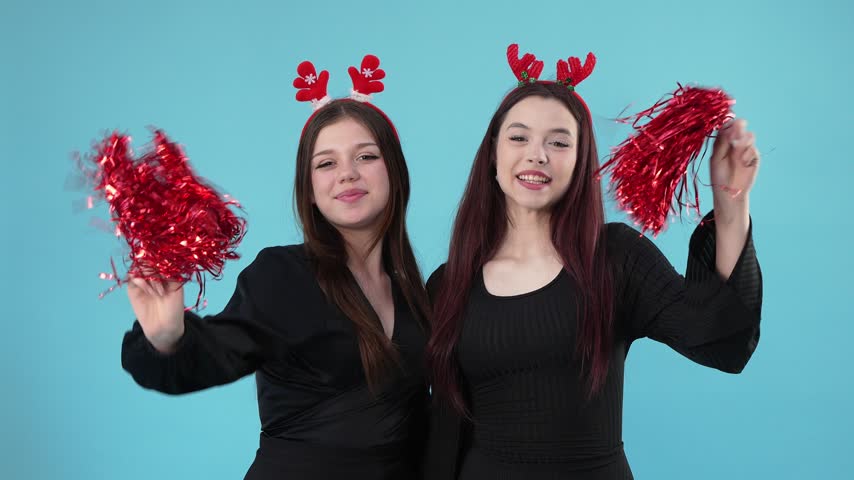 Two young women in black outfits wearing festive reindeer headbands and holding red pom-poms while posing against a blue background.