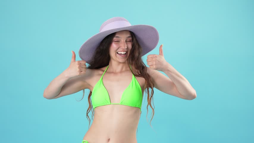 A smiling young woman in a green bikini wearing a wide-brimmed purple sun hat and showing thumbs up.