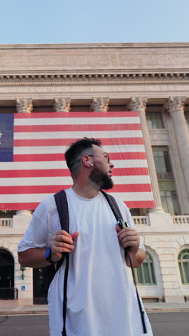 Vertical Screen: Washington DC tourist with beard and glasses wearing white t-shirt and backpack stands in front of Department of Agriculture building with large American flag during golden hour.