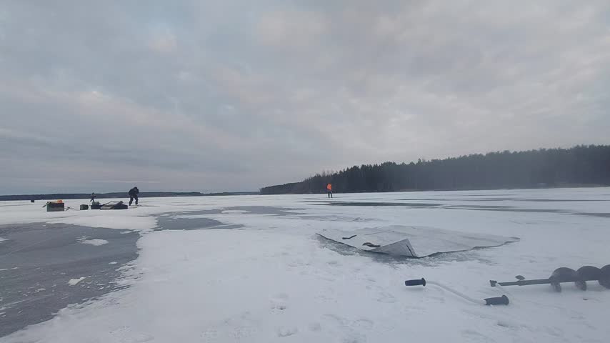People are ice fishing on a frozen lake during winter. They are set up on the snow, working with their gear. In the background, trees give a view of the landscape. Some individuals are focused on their tasks while others are scattered around the lake.