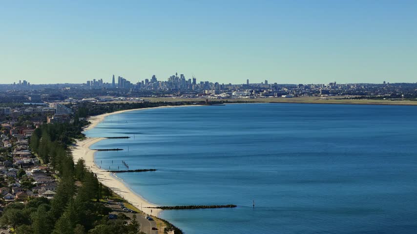 Brighton-Le-Sands beach curving toward Sydney skyline across Botany Bay, aerial panoramic establishing orbit
