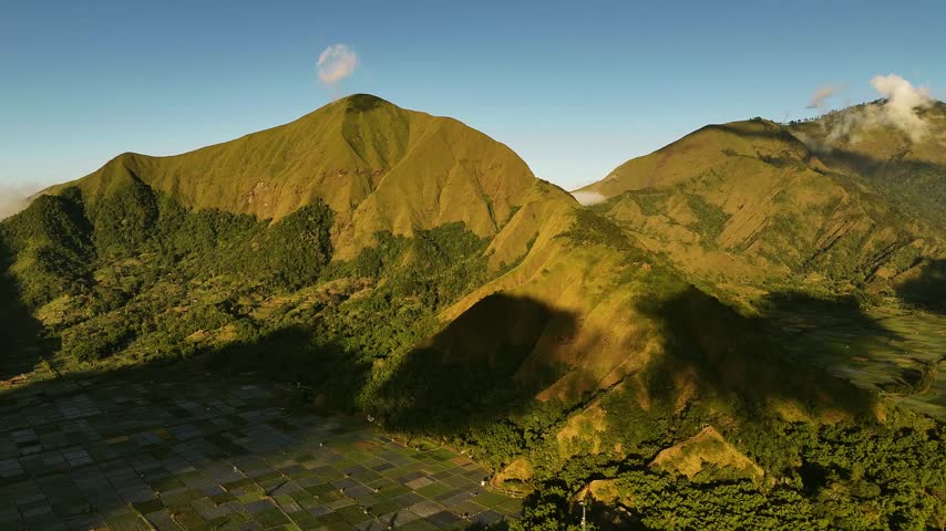 Sembalun Valley beneath lush volcanic peaks in warm morning light, aerial panoramic establishing overview above geometric farm plots