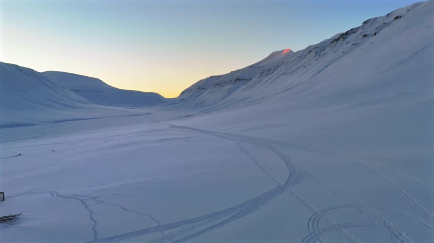 A sled dog team and musher depart a kennel yard lined with dog houses, beneath a distant peak glowing with sunrise light in Svalbard, Norway, drone pullback