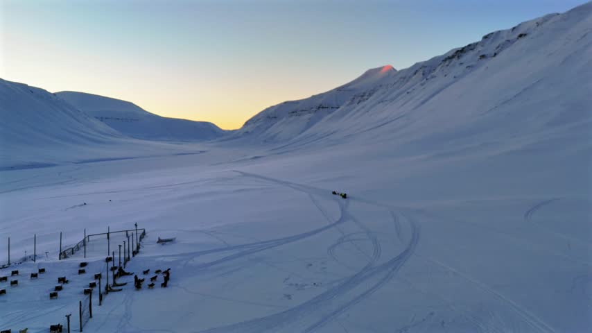 A guide sets out from the kennel lodge with a dog sled team, beginning the route across the valley as pink sunrise light touches a distant peak in Svalbard, Norway, static shot