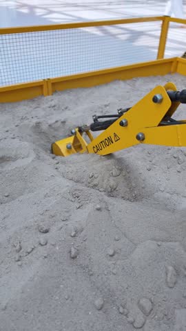A close-up vertical shot of a yellow miniature toy excavator bucket digging and scooping loose sand. The mechanical arm operates inside a fenced indoor sandbox play area at a brightly lit shopping mal