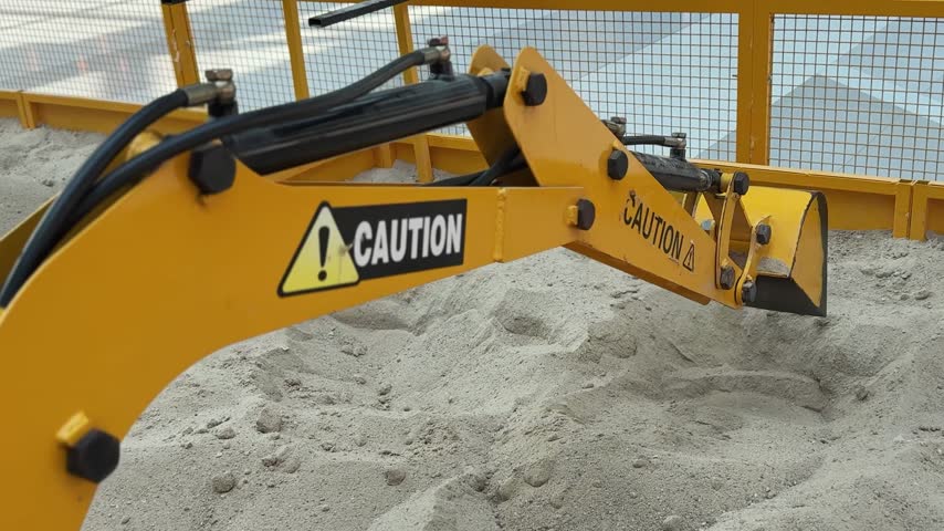 A close-up shot of a yellow miniature toy excavator bucket pouring sand into a pile. The mechanical digger with caution stickers operates inside a bright enclosed indoor play area sandbox.