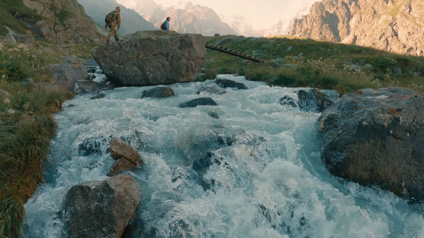 Two hikers walk across a wooden bridge over a rocky stream in an alpine valley during golden hour. The turquoise water flows over stones in warm sunlight. Snow-capped peaks rise beyond green slopes