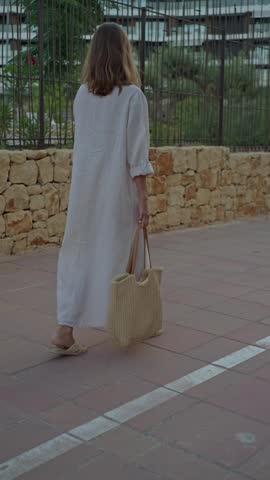 A person walks on a stone pathway while holding a bag. The surroundings show a simple landscape with a stone wall and some greenery. The time appears to be during the day.