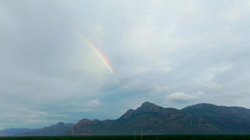 Rainbow arcs through storm clouds over rugged mountains in India, dense coconut plantation below, dramatic monsoon light, cinematic drone shot of scenic Indian countryside landscape.
