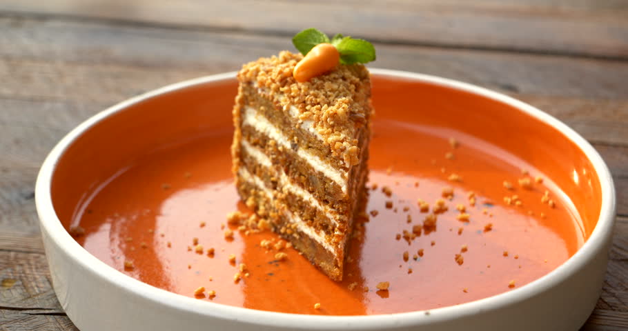 Hand presses a fork into a tall layered carrot cake then lifts a tender bite revealing alternating moist sponge and cream filling beneath nut crumbs mint leaves and a small decorative carrot