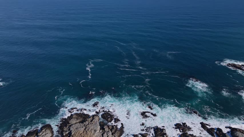 Retreating flight over a rocky shoreline with crashing waves and a coastal road revealing scattered clifftop houses along the deep blue sea
