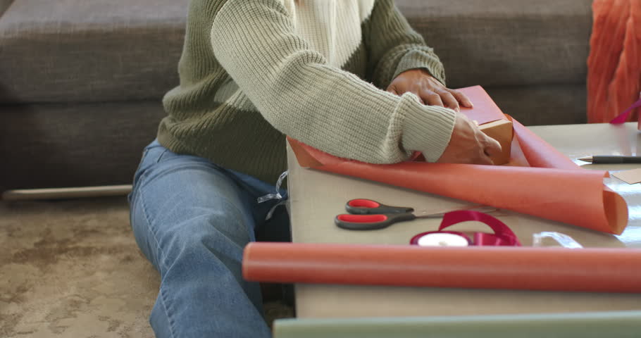 African American woman placing box wrapping orange paper making gift at home wearing olive sweater. Present, folding, tape, ribbon, scissors, hands, sitting