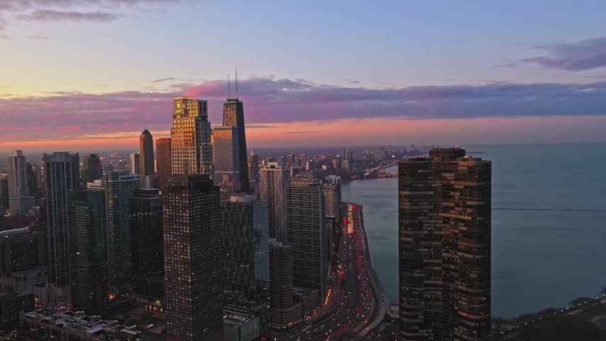 Aerial footage moves forward over downtown Chicago at sunset, revealing skyscrapers, Lake Michigan and busy lakeshore traffic beneath a colorful pink, purple and orange evening sky.