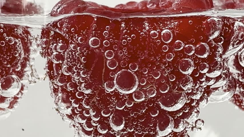 Bubbles surrounding red raspberries in sparkling water. The bubbles move around the raspberries.